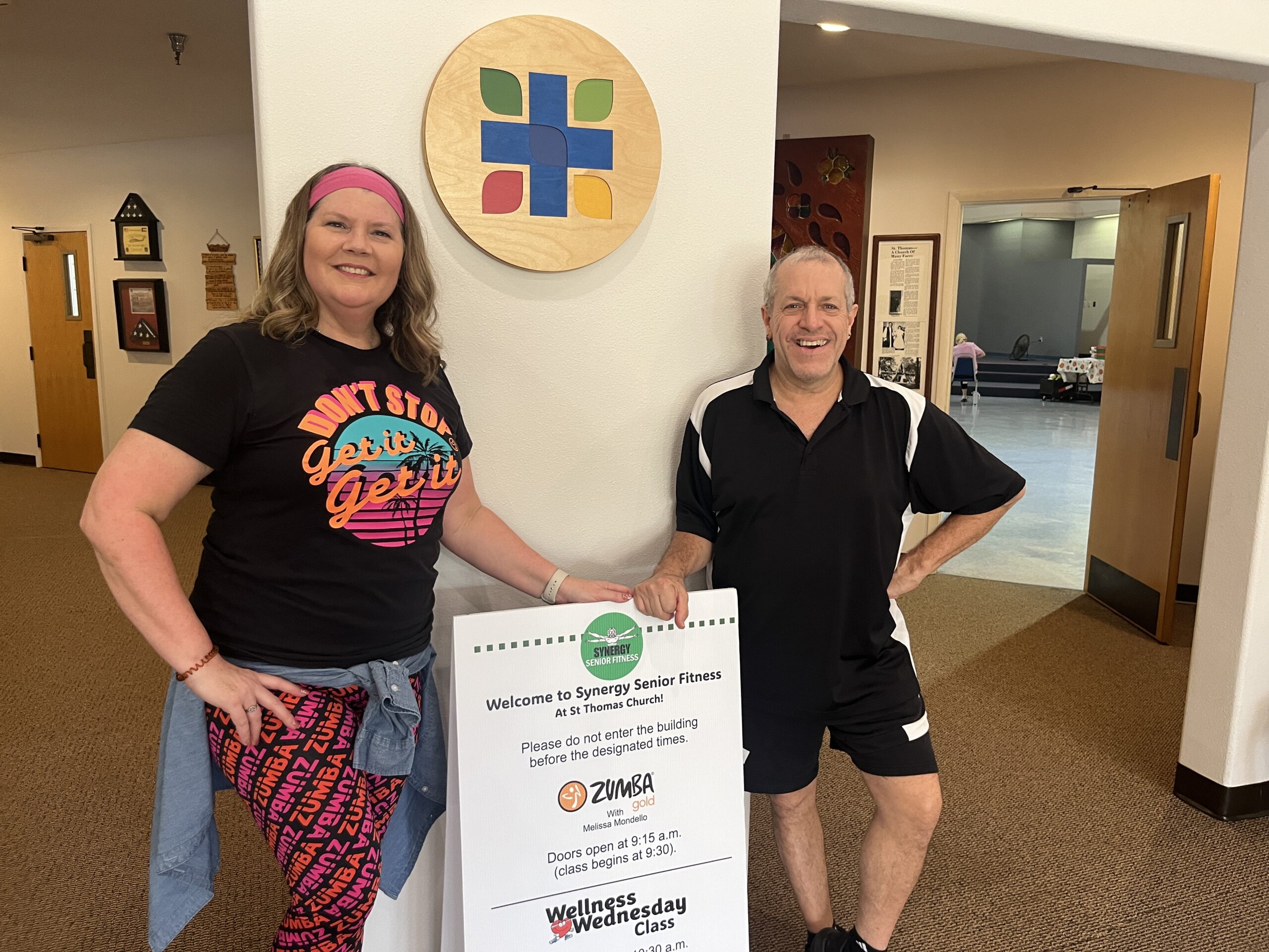 Two people stand smiling in a fitness center hallway One wears colorful exercise attire and a headband, and the other is in black workout clothes They are next to a sign for a senior fitness class A round logo hangs on the wall behind them