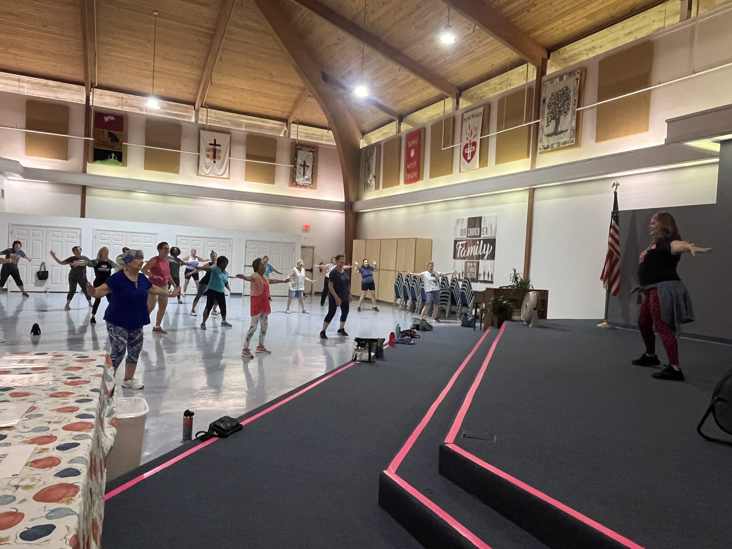 A group of people participates in a dance class led by an instructor in a large hall with high wooden ceilings The room has an American flag and decorative banners on the walls