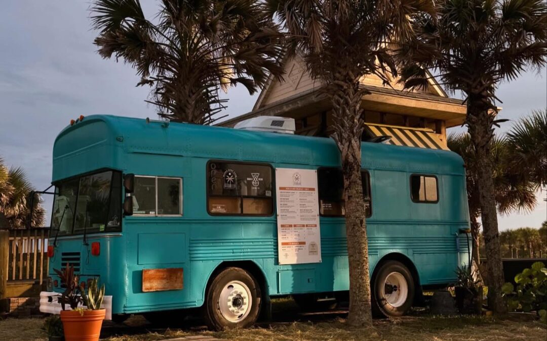 A turquoise bus converted into a café is parked under palm trees It has a counter window on one side with a menu displayed The setting appears tropical with grass and a few plants surrounding the area