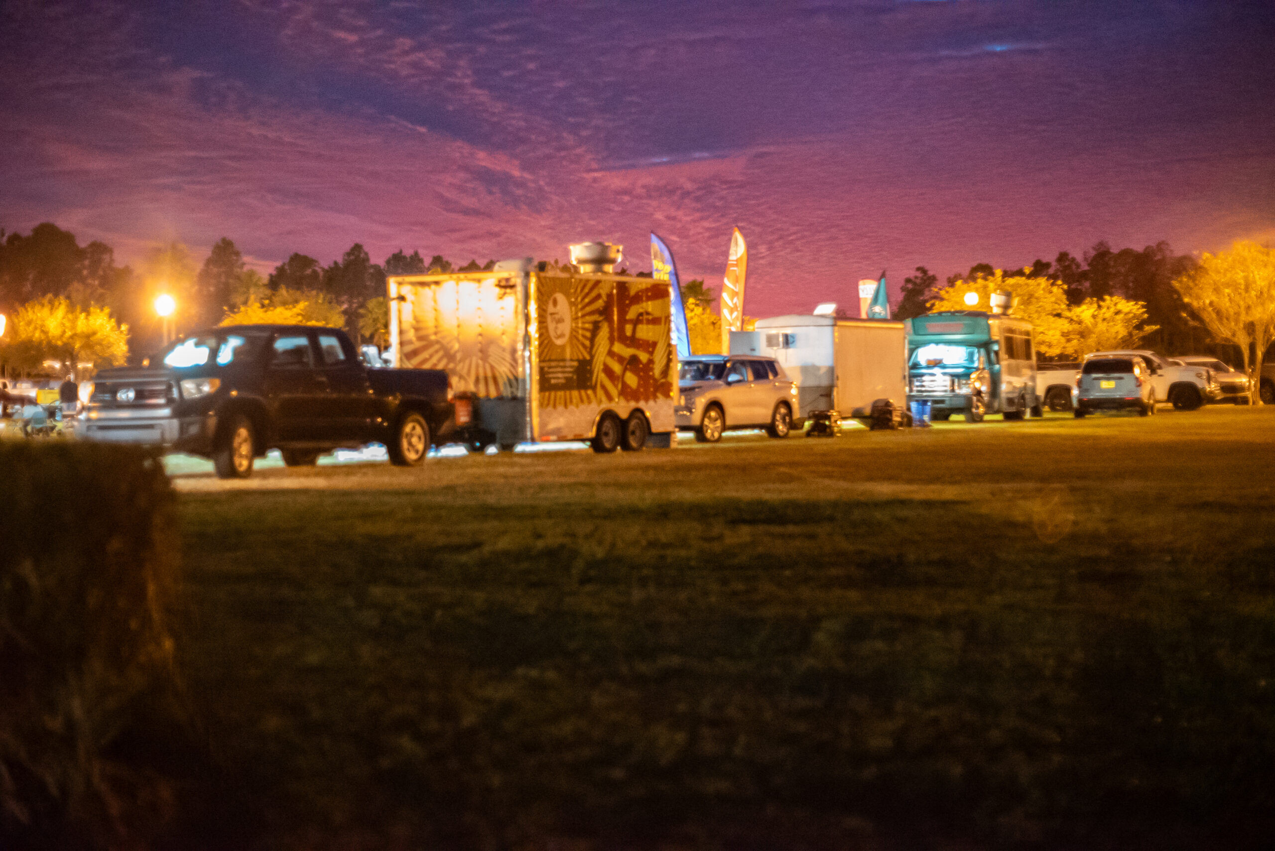 A row of colorful food trucks and parked cars under a vibrant purple and orange evening sky Trees and warm streetlights create a cozy atmosphere at the outdoor event