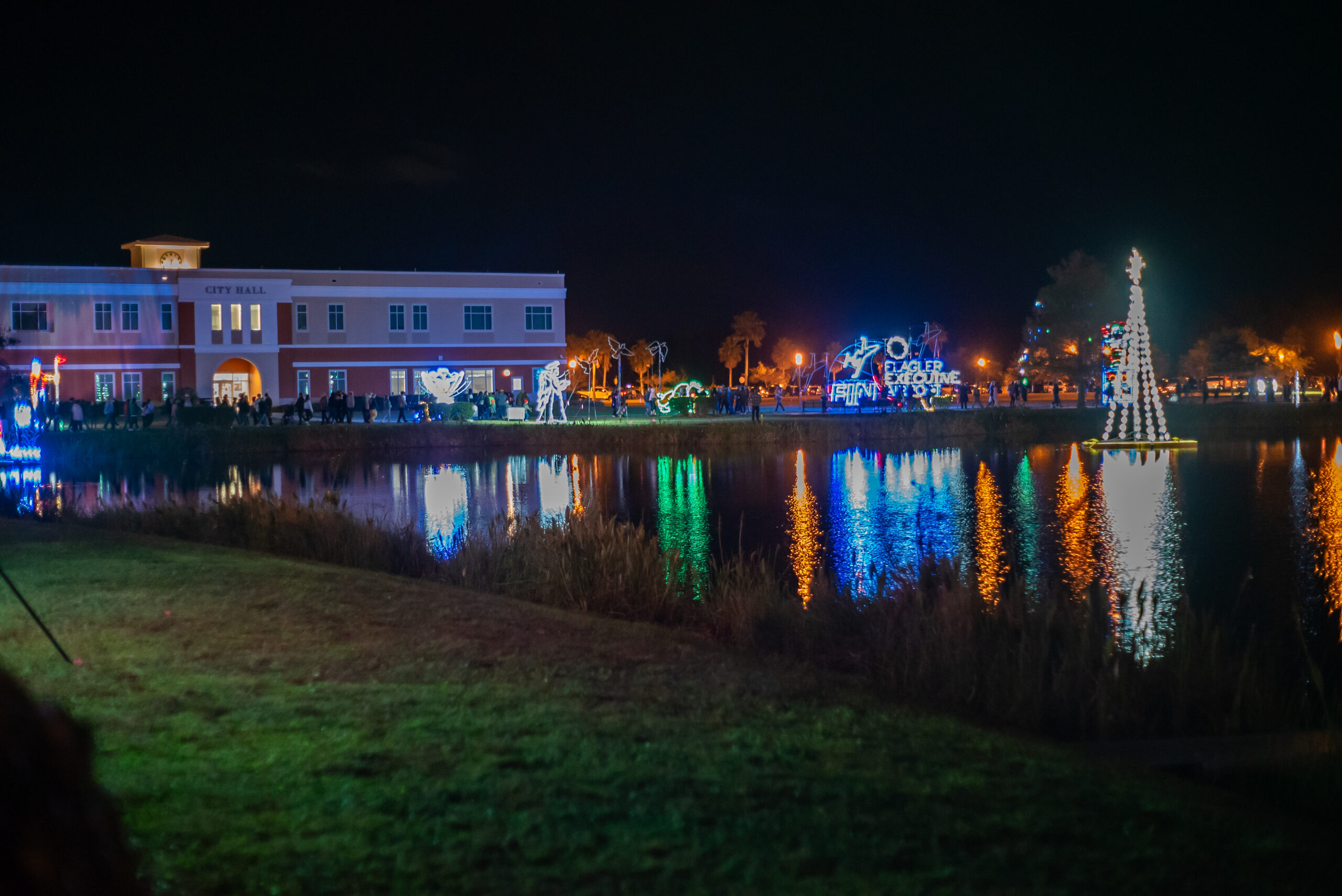A nighttime scene by a lake, featuring festive holiday lights reflecting on the water The decorations include illuminated figures and a tall tree In the background, a building marked "City Hall" is visible, with people gathered nearby