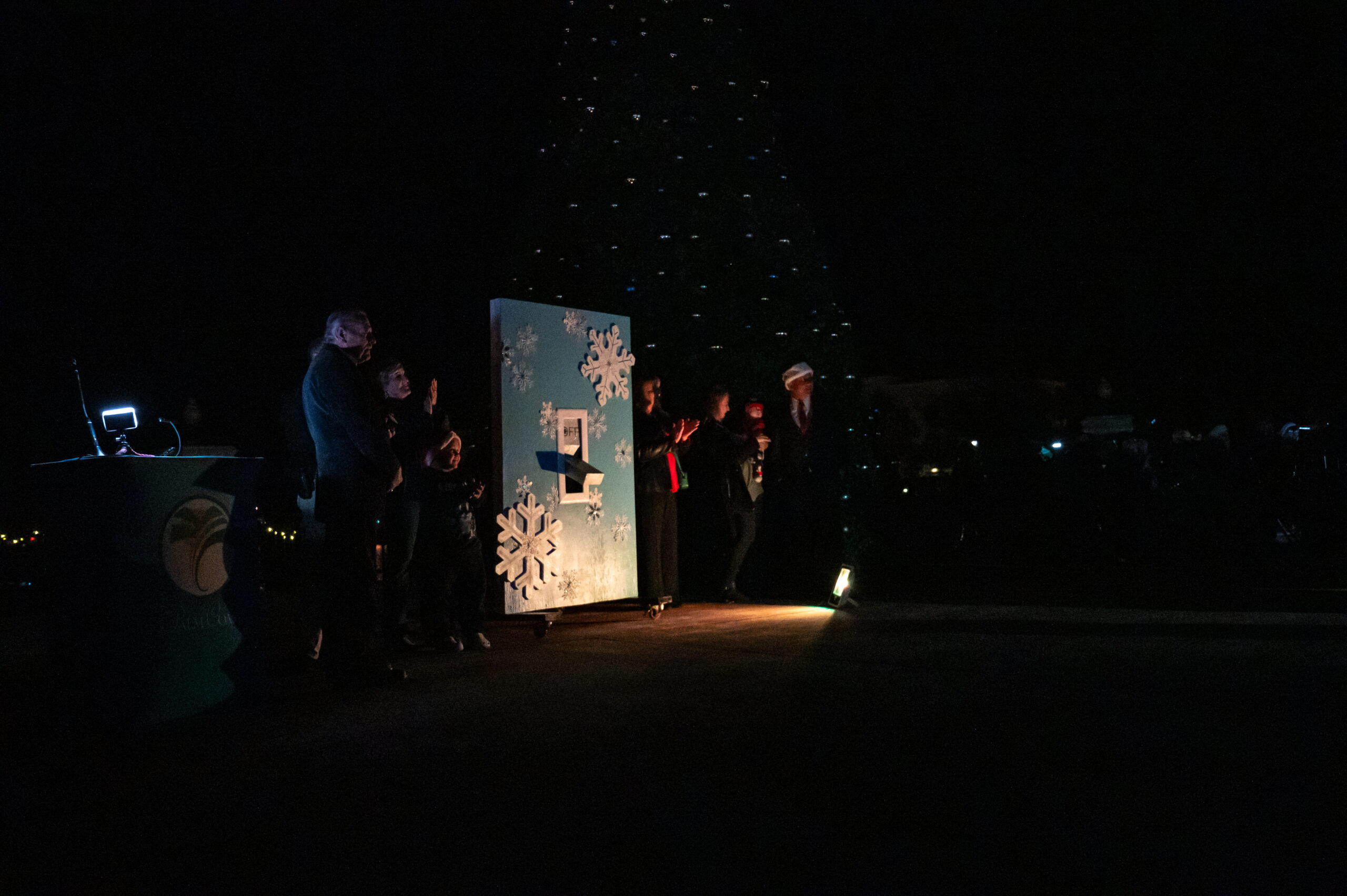 A dark outdoor scene showing a group of people next to a large board decorated with snowflakes A dimly lit Christmas tree is partially visible in the background A small table with a light fixture is on the left