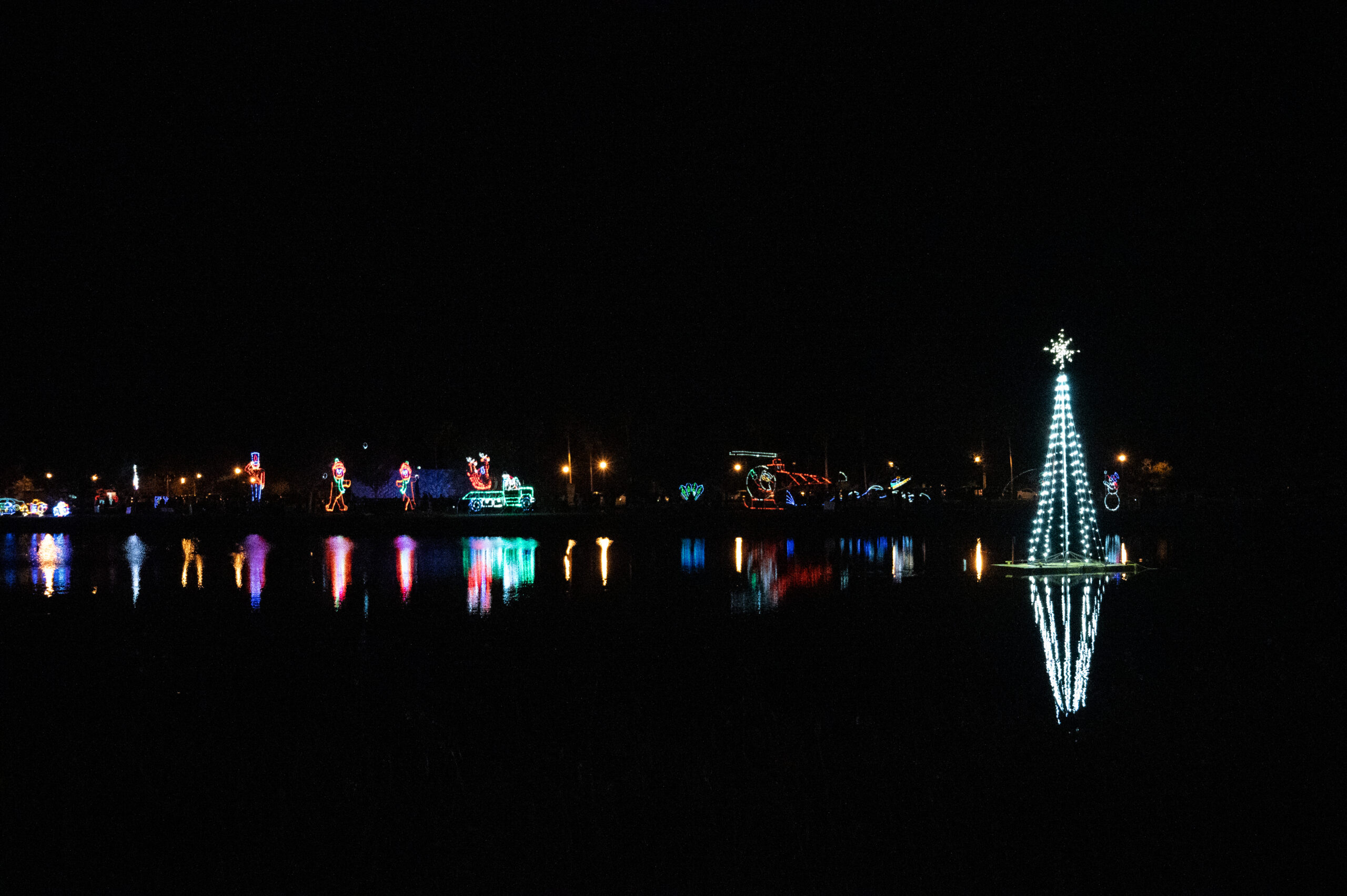 A serene night scene featuring colorful holiday lights reflecting off a calm body of water On the right, a large, illuminated tree shaped display stands tall, its reflection shimmering on the surface below