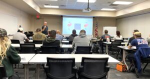 A classroom with several adults seated at tables, facing a teacher giving a presentation The screen displays a diagram, and an American flag is in the corner The atmosphere is focused and educational