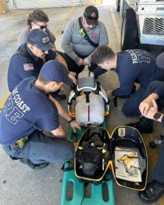 A group of firefighters and emergency responders gather around medical equipment in a garage They are focused on a device labeled "LUCAS " and examining a kit on the ground They wear uniforms with "PALM COAST FIRE" printed on the back