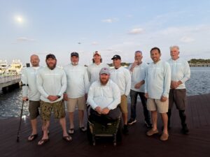 A group of nine men, wearing light blue shirts, stand and sit on a wooden dock by the water One man in a wheelchair is in front They all appear to be relaxed and smiling, with boats and a partly cloudy sky in the background