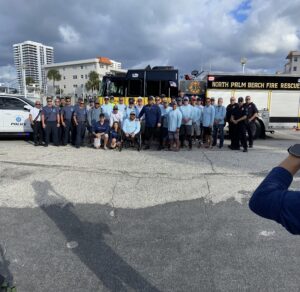 A group of people, including firefighters and police officers, pose in front of a fire engine and a police car The backdrop features buildings and a partly cloudy sky