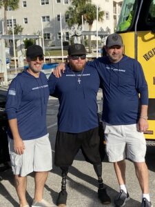 Three men wearing matching "Hookin' Veterans" shirts and hats stand outdoors on a sunny day The man in the middle has prosthetic legs They smile and pose together casually in front of a yellow vehicle