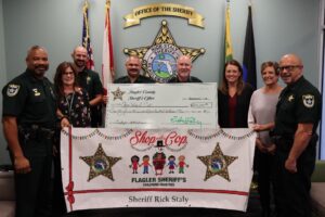 A group of people, including law enforcement officers and civilians, stand together holding a large ceremonial check and a banner for Flagler Sheriff's Children's Charities Flags and an official emblem are displayed in the background
