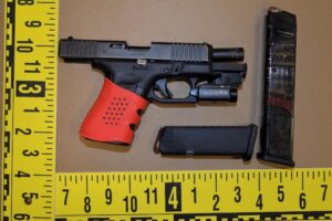 A black handgun with a red grip and attached flashlight sits next to a black magazine on a brown surface, surrounded by a yellow measuring tape with black markings indicating inches
