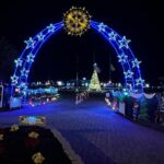 A nighttime scene features a large arch adorned with blue and white lights, topped by a star shaped decoration In the background, a brightly lit Christmas tree stands, surrounded by festive lights and decorations A paver path leads under the arch
