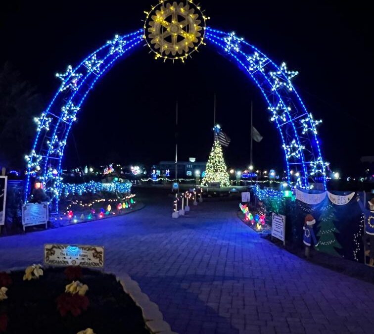 A nighttime scene features a large arch adorned with blue and white lights, topped by a star shaped decoration In the background, a brightly lit Christmas tree stands, surrounded by festive lights and decorations A paver path leads under the arch