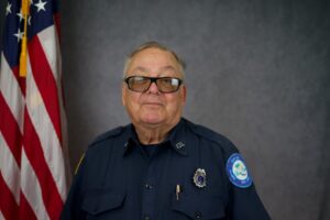 An older person wearing a dark blue uniform poses for a portrait, standing in front of an American flag The uniform has various patches and insignia on the sleeves and chest The background is a neutral gray