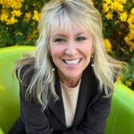 A smiling woman with long blonde hair is sitting in a green chair She is wearing a black blazer over a beige top Behind her, there is a backdrop of bright yellow flowers
