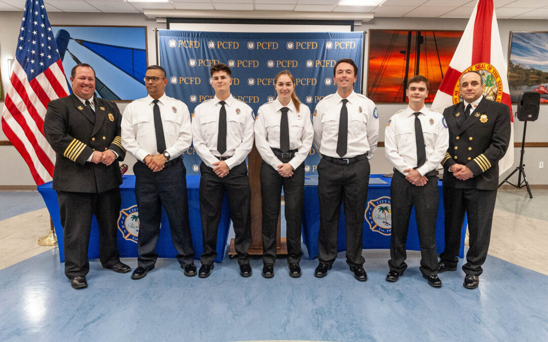 A group of seven people, including six in white shirts and ties and one in a dark uniform, stand smiling in a row indoors with a backdrop featuring PCFD logos American and other flags are visible on each side
