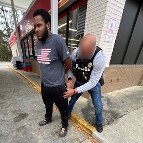 A person is being handcuffed by a police officer outside a building The officer is wearing a vest labeled "POLICE," and the person being handcuffed is wearing a t shirt and pants The setting appears to be near a convenience store
