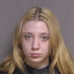 A person with long, light blonde hair is looking directly at the camera with a neutral expression The background is plain and gray