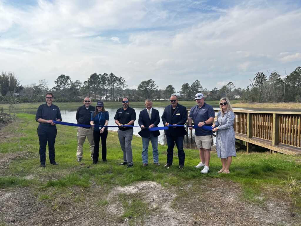 A group of eight people standing outdoors, all holding a long blue ribbon ready to be cut They are in a grassy area near a wooden deck with trees and a pond in the background The sky is cloudy