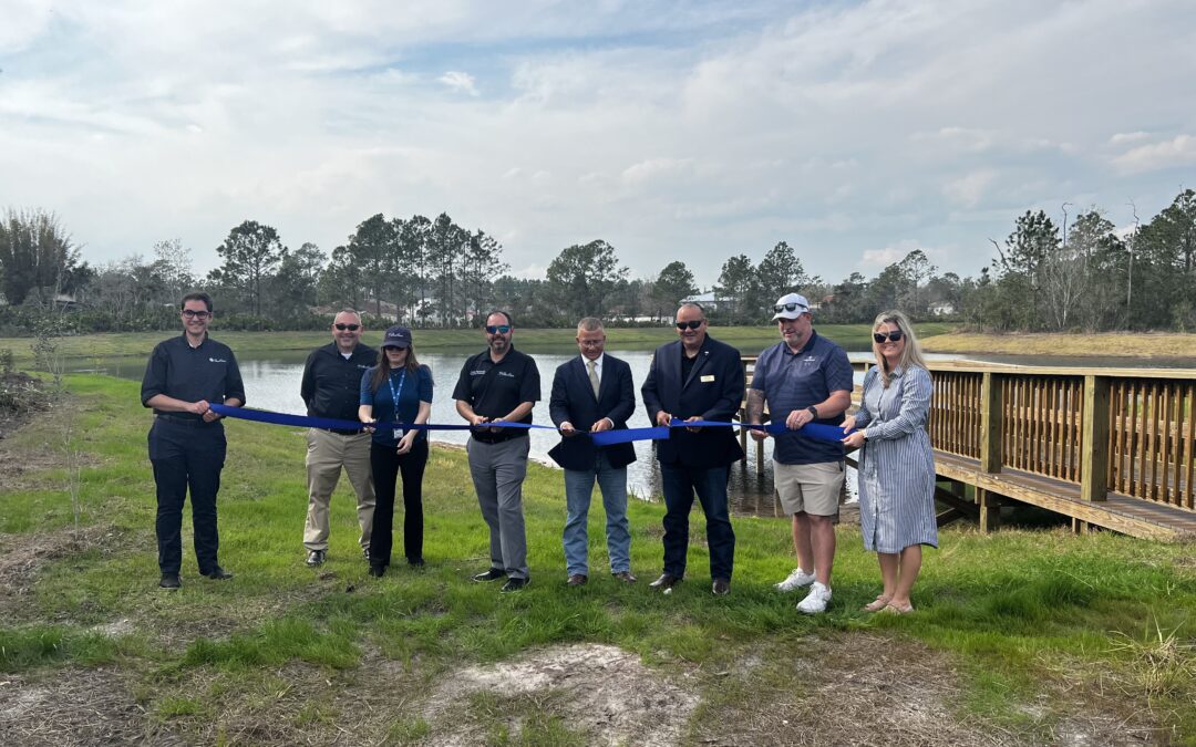 A group of eight people standing outdoors, all holding a long blue ribbon ready to be cut They are in a grassy area near a wooden deck with trees and a pond in the background The sky is cloudy