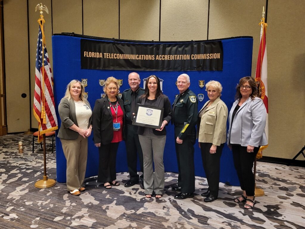 Seven people stand in front of a Florida Telecommunications Accreditation Commission banner The central woman holds a certificate They are dressed in formal and uniform attire, and the setting includes two flags on either side