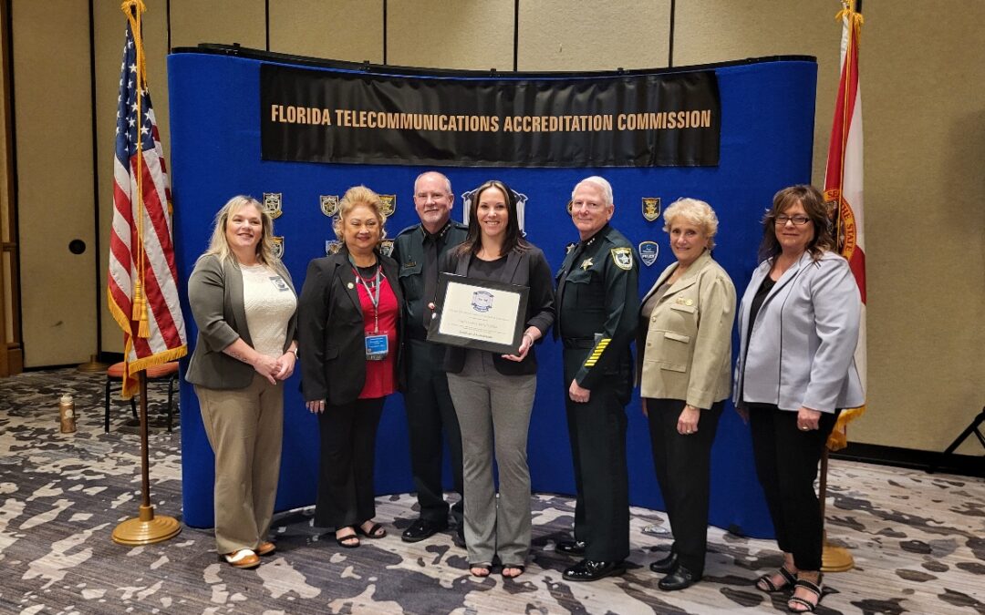 Seven people stand in front of a Florida Telecommunications Accreditation Commission banner The central woman holds a certificate They are dressed in formal and uniform attire, and the setting includes two flags on either side