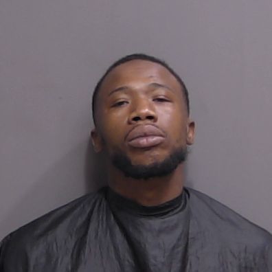 A man with short hair and a trimmed beard wearing a black drape poses against a plain gray background for a mugshot