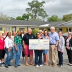 A diverse group of adults stands outside a yellow building, smiling and gathered around a large ceremonial check for $, made out to Fischer For Care Trees and a cloudy sky are in the background