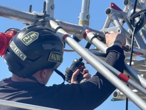 A person wearing a helmet labeled "Pollock" uses a power tool to secure metal scaffolding under a clear blue sky