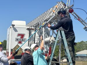 Three men wearing helmets work together on a ladder truck One man stands on a ladder adjusting equipment, while the others assist from the ground The ladder truck is labeled "MARC " It’s a sunny day