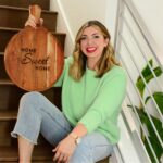 A woman with blonde hair, wearing a light green sweater and jeans, sits on stairs holding a round wooden board engraved with the words "Home Sweet Home " She is smiling and has bright red lipstick