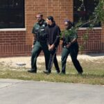 Two police officers escort a handcuffed person in black clothing walking on a sidewalk beside a brick building and some grass under a sunny sky