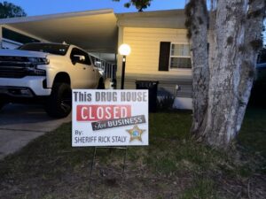 A house with a truck parked in the driveway A sign in the yard reads, "This DRUG HOUSE CLOSED FOR BUSINESS by: SHERIFF RICK STALY," with a sheriff's badge graphic