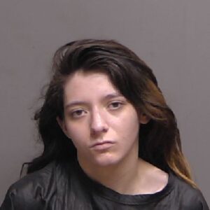 A young woman with long dark hair, wearing a dark top, looks at the camera with a neutral or serious expression against a plain background