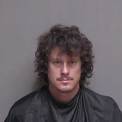 A person with curly brown hair and a mustache is wearing a black drape and sitting against a plain gray background