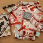 Assorted evidence bags and tubes labeled with red tape, containing various items, surround a handgun, two magazines, and loose bullets arranged in a white box A chalkboard labeled "WU TANG" is visible among the bags