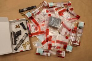 Assorted evidence bags and tubes labeled with red tape, containing various items, surround a handgun, two magazines, and loose bullets arranged in a white box A chalkboard labeled "WU TANG" is visible among the bags