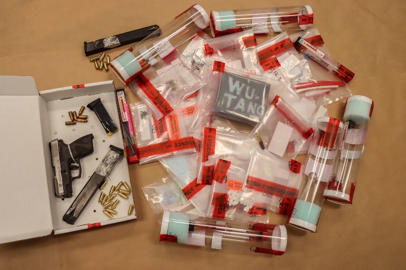 Assorted evidence bags and tubes labeled with red tape, containing various items, surround a handgun, two magazines, and loose bullets arranged in a white box A chalkboard labeled "WU TANG" is visible among the bags