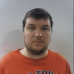 A man with short dark hair and light facial hair, wearing a red T shirt, standing against a plain light background, facing the camera with a neutral expression
