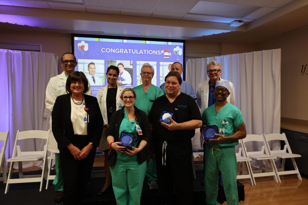 A group of ten medical professionals poses indoors, some in scrubs and others in business attire, holding blue glass awards Behind them is a screen displaying "CONGRATULATIONS" and photos, with white curtains and empty chairs