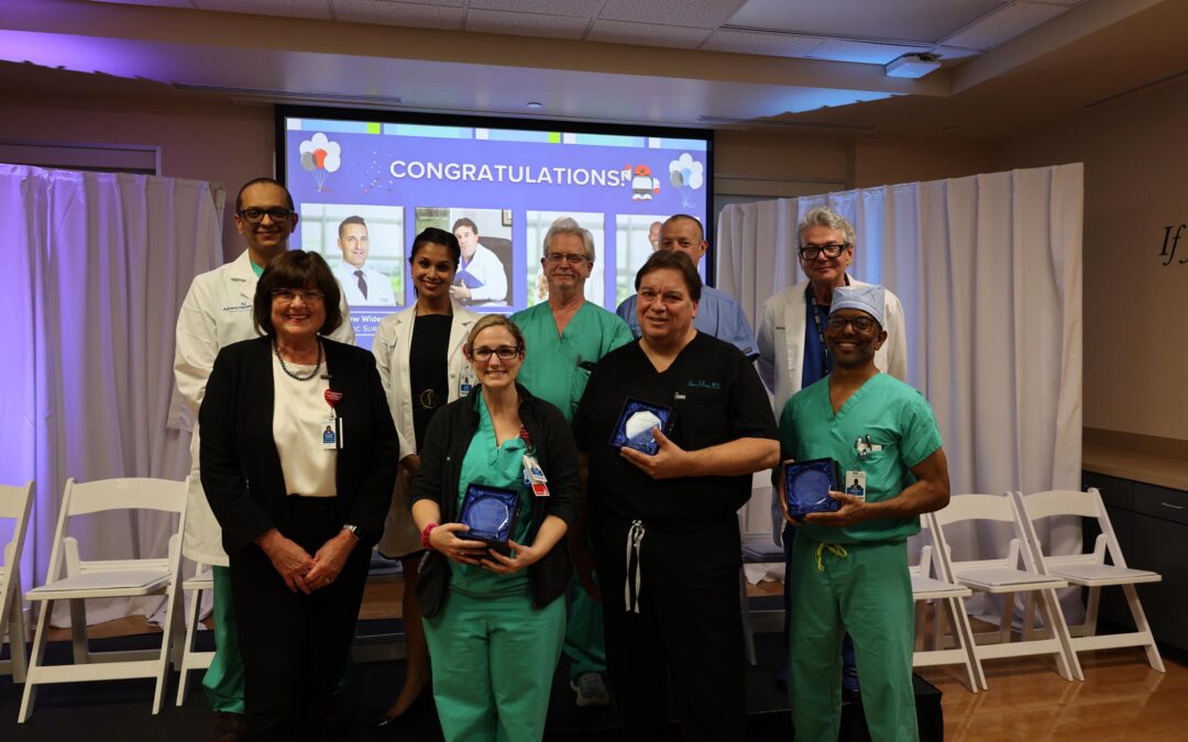 A group of ten medical professionals poses indoors, some in scrubs and others in business attire, holding blue glass awards Behind them is a screen displaying "CONGRATULATIONS" and photos, with white curtains and empty chairs