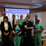 A group of ten medical professionals poses indoors, some in scrubs and others in business attire, holding blue glass awards Behind them is a screen displaying "CONGRATULATIONS" and photos, with white curtains and empty chairs