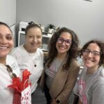 Four women smiling inside an office; one holds a gift bag with red tissue paper They wear casual clothes, two in printed t shirts, one in a blazer, and appear cheerful and friendly Office supplies and plants are visible in the background