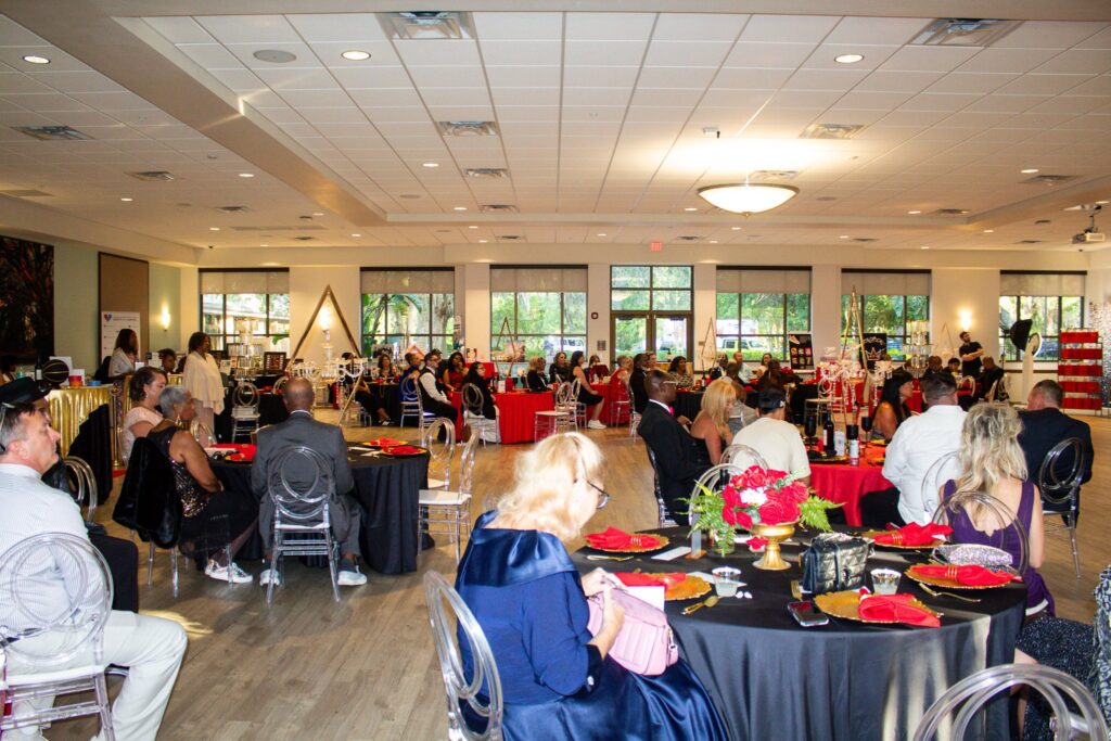 A formal event with guests seated at round tables covered in black and red tablecloths, decorated with red floral centerpieces, in a brightly lit room with large windows and a wooden floor