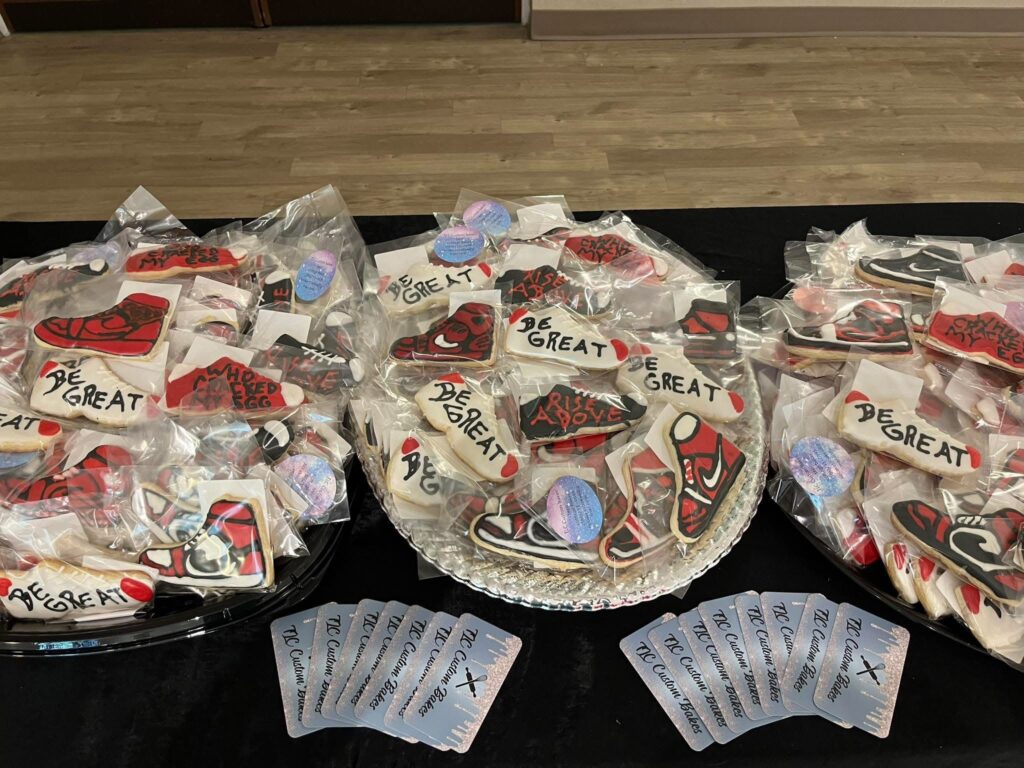 Decorated cookies shaped like red and black sneakers and plaques with phrases like “BE GREAT” and “RISE ABOVE” are displayed on trays and in bags Nearby are multiple clear plastic cards labeled “TLC Cookie Creations ”