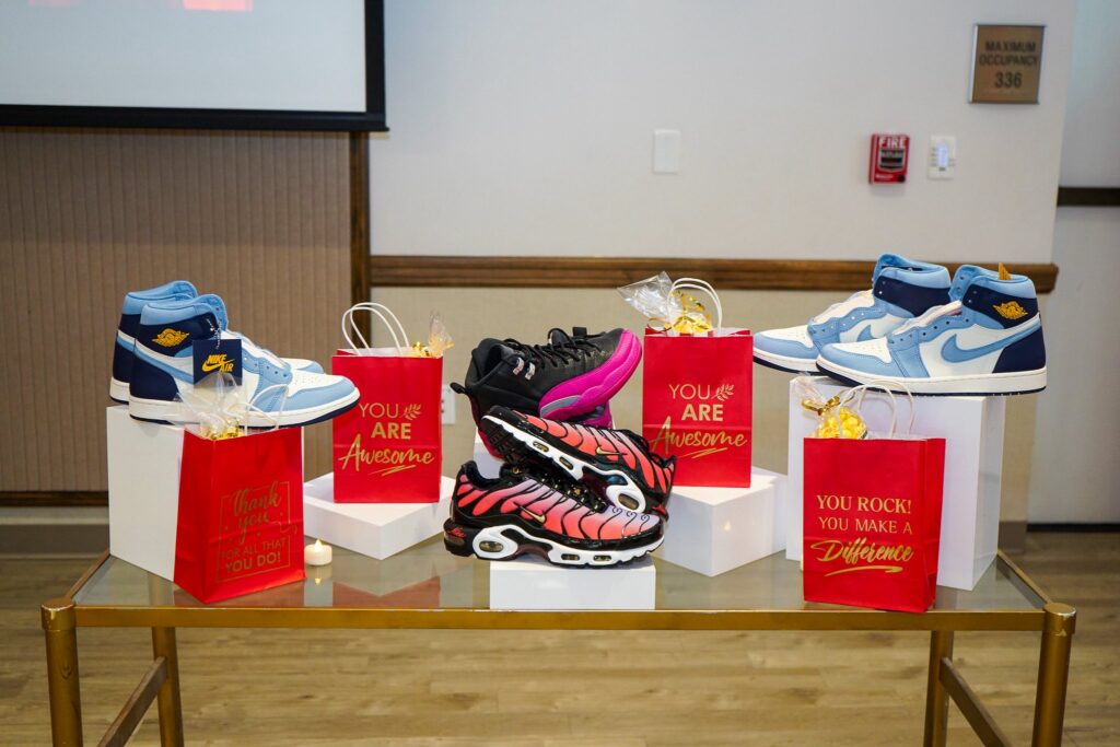 A display of four pairs of colorful sneakers and four red gift bags with uplifting messages on a glass table, set against a beige wall in a room Each gift bag has gold ribbon and a small gift inside A display of four pairs of colorful sneakers and four red gift bags with uplifting messages on a glass table, set against a beige wall in a room Each gift bag has gold ribbon and a small gift inside