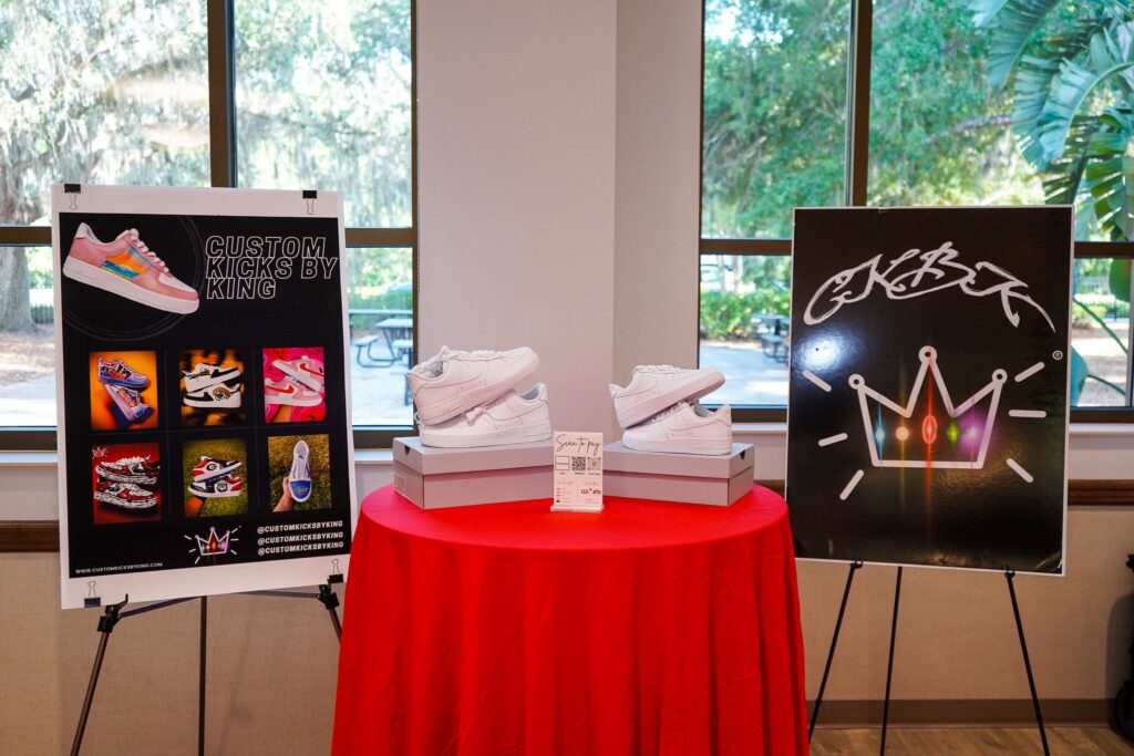 A display table with two pairs of white sneakers sits between two posters—one shows various colorful custom sneakers, the other features a graffiti style crown—set in front of large windows with greenery outside A display table with two pairs of white sneakers sits between two posters—one shows various colorful custom sneakers, the other features a graffiti style crown—set in front of large windows with greenery outside
