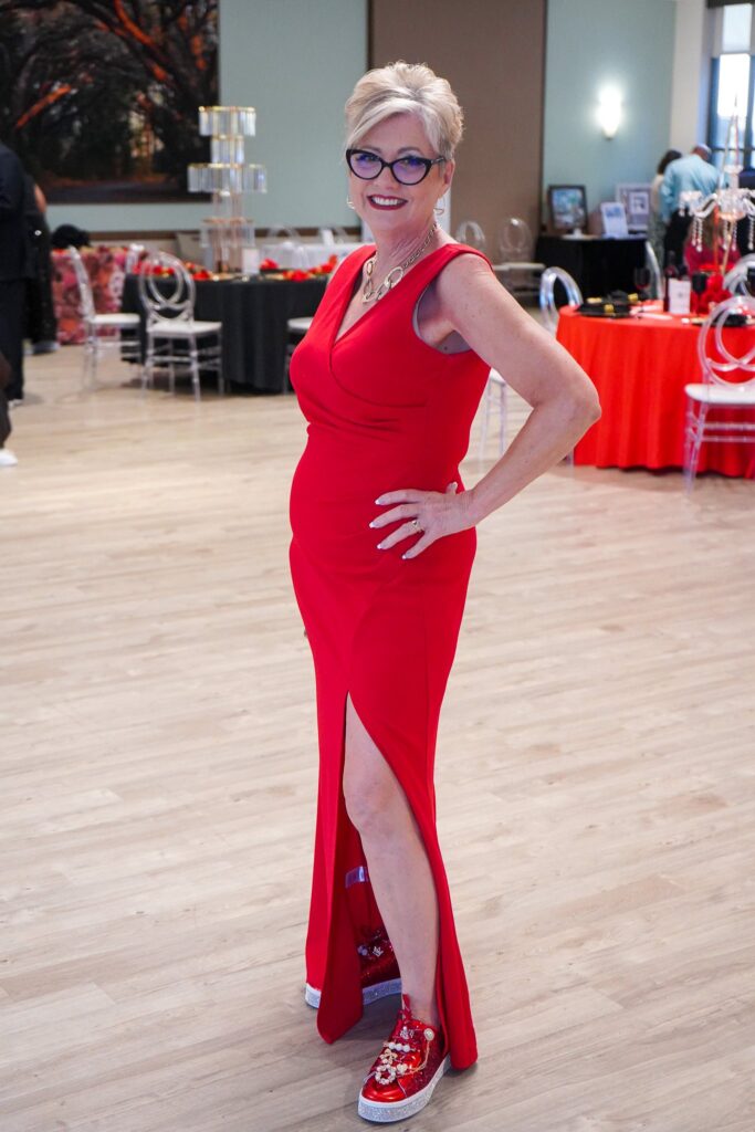 A woman in a sleeveless red gown with a thigh high slit poses indoors, smiling She wears glasses, a necklace, and red sneakers with floral designs The background shows tables and chairs in a spacious event room A woman in a sleeveless red gown with a thigh high slit poses indoors, smiling She wears glasses, a necklace, and red sneakers with floral designs The background shows tables and chairs in a spacious event room