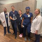 Five healthcare workers in scrubs and lab coats stand smiling in front of a row of lockers One person holds an award The setting appears to be a hospital or clinic staff room