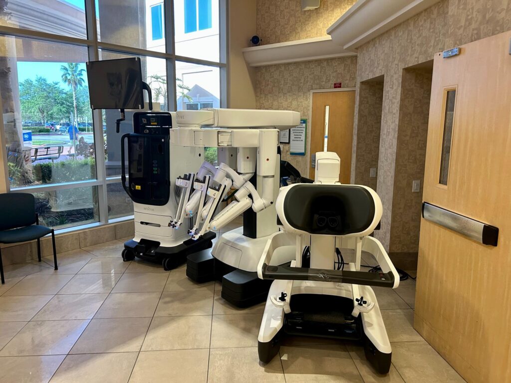Two advanced robotic surgery machines are positioned in a hospital lobby with large windows, beige walls, and tiled floors Trees and a courtyard are visible outside through the windows