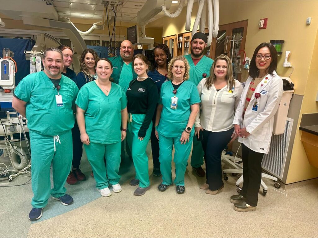 A group of healthcare professionals, some in green scrubs and others in business attire, pose and smile together in a hospital or medical facility Medical equipment is visible in the background